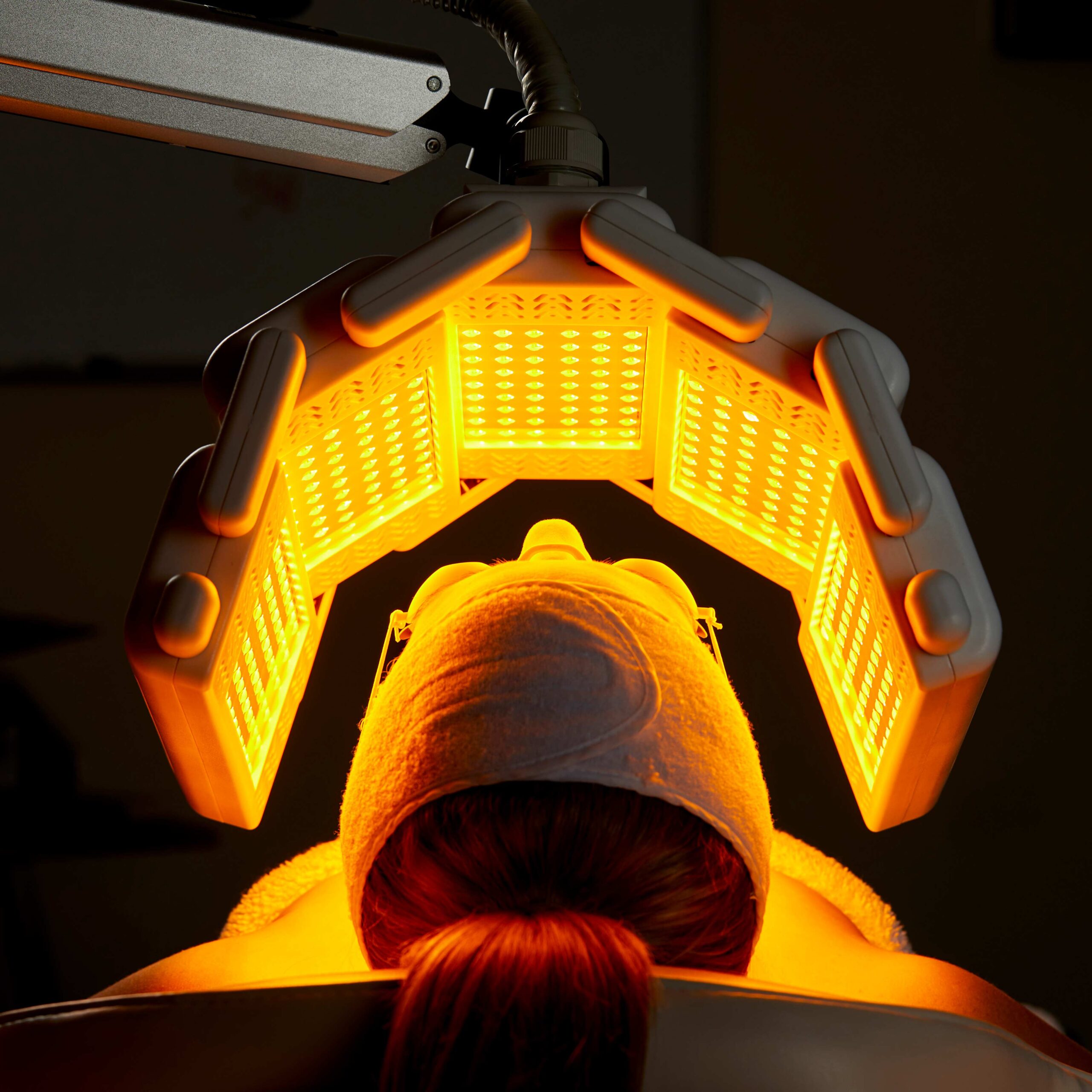 Woman lies on a bed and has a beauty machine over her face, shining LED lights for a skin treatment
