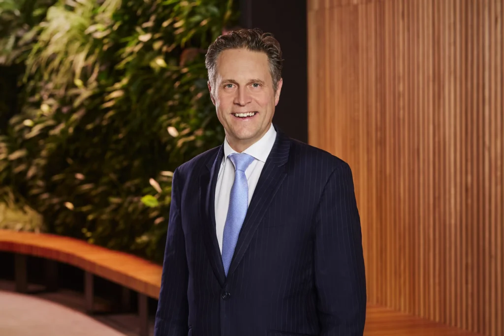 Personal branding headshot of a smiling man wearing a navy suit and light blue tie against a living green wall backdrop.