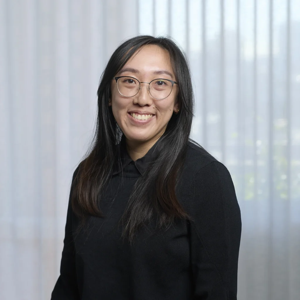 Team consistency headshot of a smiling woman with glasses and long dark hair in a black top, posed against a uniform white sheer curtain backdrop.