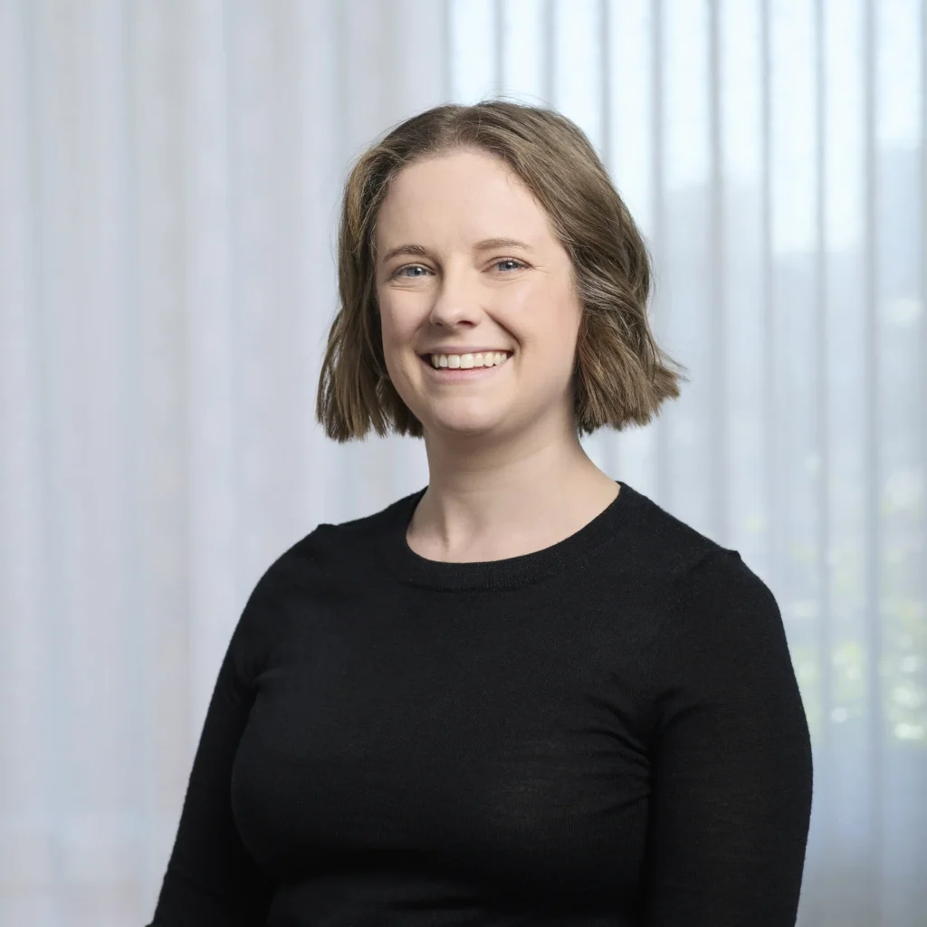 Team consistency headshot of a smiling woman with short wavy hair wearing a black long-sleeve top, posed against a uniform white sheer curtain backdrop.