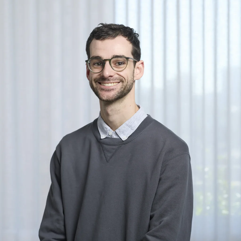 Team consistency headshot of a smiling man with glasses and a short beard wearing a grey crewneck sweater, posed against a uniform white sheer curtain backdrop.