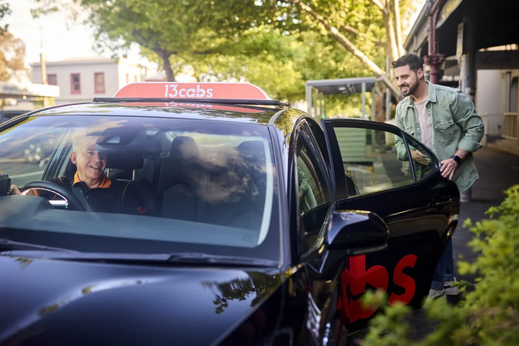 Lifestyle commercial photography showing a male passenger opening the door of a 13cabs vehicle while the driver smiles from inside.