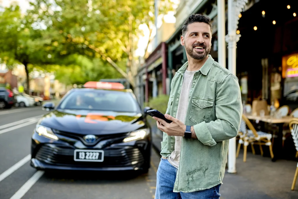 On-location commercial photography of a man holding a smartphone on a Melbourne sidewalk with a black 13cabs taxi parked in the background.