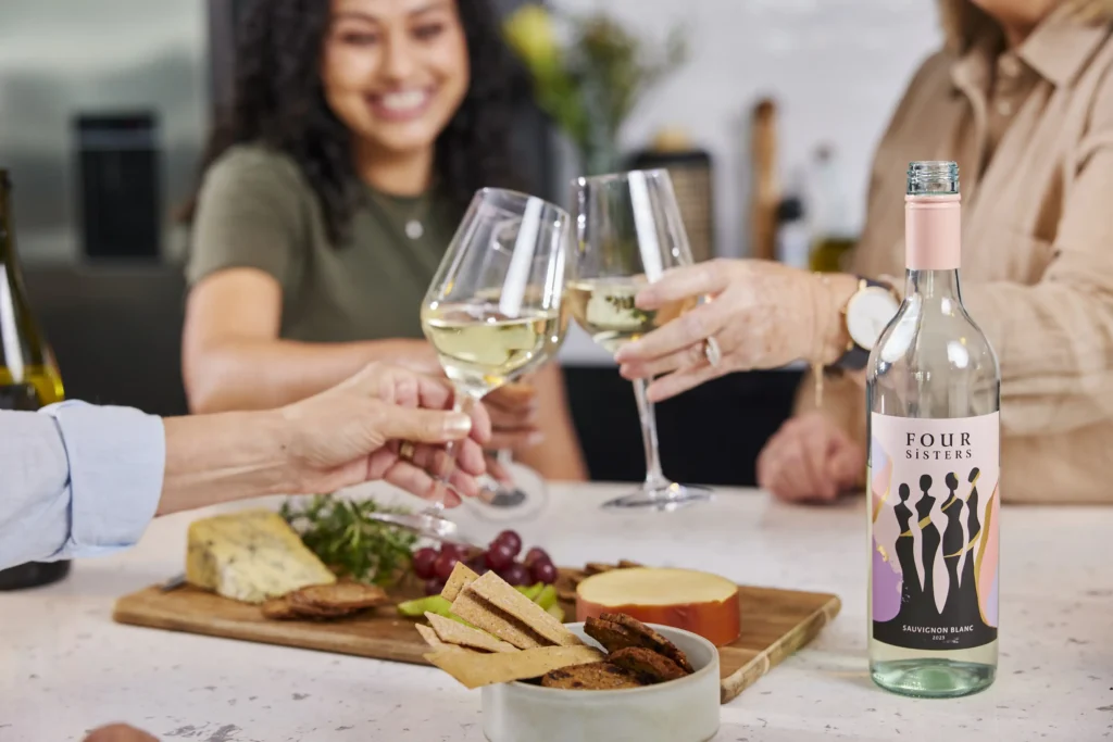 Women smiling and chatting around a beautifully styled, sunlit dining table with wine glasses, taken for the 4 Sisters brand.